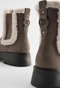 Bruine enkellaarzen met een nep-fur afwerking, zilveren hardware-accenten en een gestructureerde zwarte rubberenzool. Gladde suede-achtige stof.