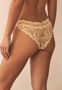 Lace hipster ondergoed in zacht beige met bloemmotieven, gegolfde randen en een doorschijnende textuur, die de verfijnde kantdetails accentueert.