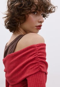 Rotes, geripptes Off-Shoulder-Oberteil mit geraffter Textur, darüber ein brauner Spaghettiträger. Nahaufnahme von Schulter und Hals, die Stoffdetails zeigen.