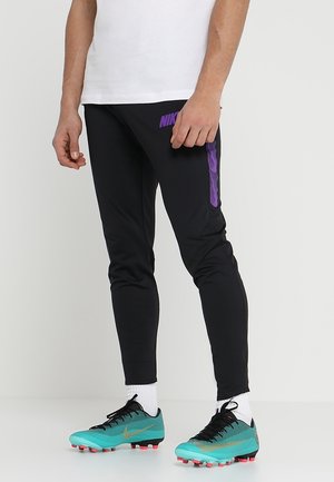 Person iført sorte Nike bukser med lilla logo og turkis fodboldstøvler med hvide sokker, stående på en hvid gulv mod en ensfarvet baggrund.