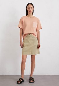 Perzikkleurig T-shirt met korte mouwen en een borstzak, gecombineerd met een beige denim rok met voorkruiszakken en een knoopsluiting. Zwarte sandalen.