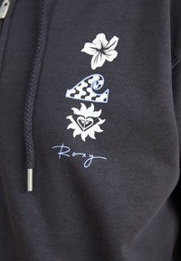 Crna majica s kapuljačom s bijelim cvjetnim i valovitim grafikonima, uz stilizirani natpis "Roxy" u svijetlo plavoj boji. Mekana, teksturirana tkanina.