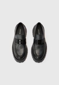 Mustat loafers, jotka on valmistettu nahasta ja synteettisistä materiaaleista, joissa on teksturoitu päällinen, leveä hihna ja paksu ulkopohja näkyvällä uralla.