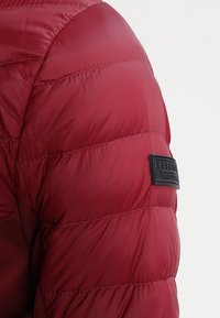 Punainen puffatakki, jossa on tikattu pinta, kiiltävä viimeistely ja musta kuminen logo-etiketti bränditunnistusta varten.