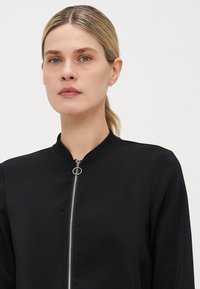 Black bomber jacket made of smooth fabric, featuring a high collar and a front zipper with a circular pull. Simple design without patterns.