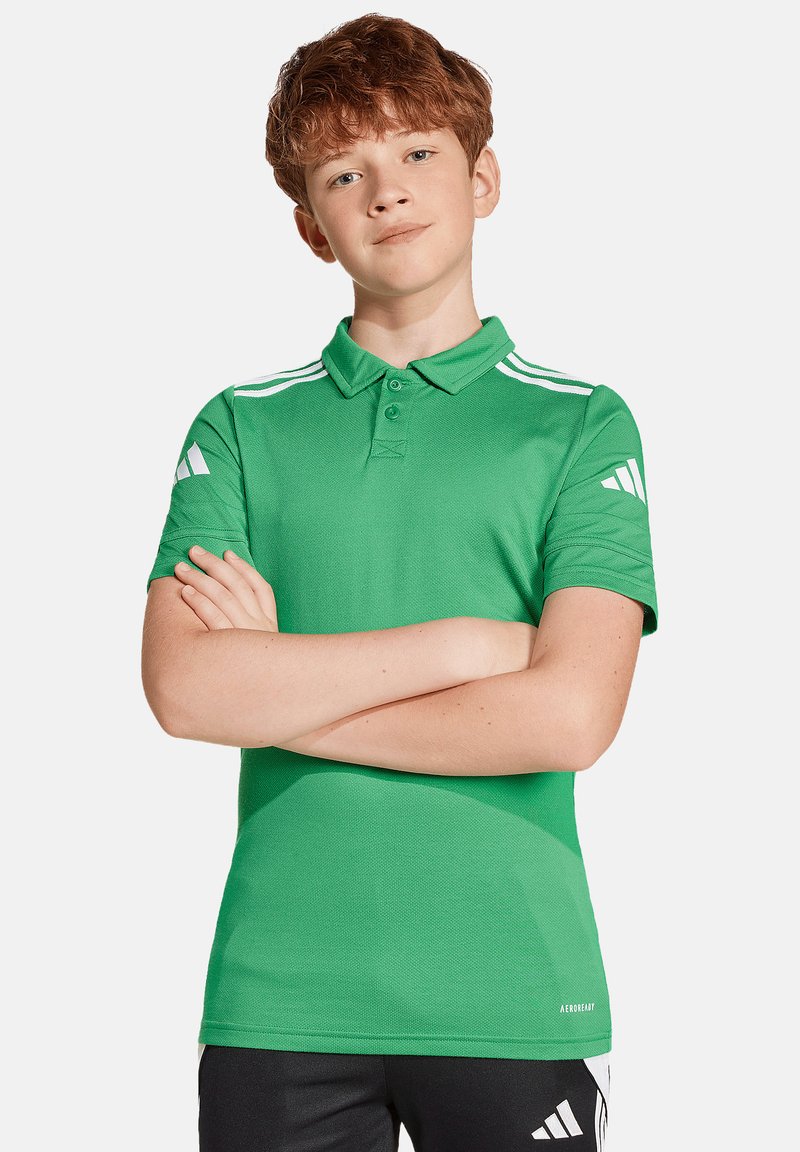 Grüne Poloshirt aus strukturiertem Stoff, mit einem Drei-Knopf-Kragen und weißen Akzenten an den Ärmeln. Kombiniert mit schwarzen Shorts.