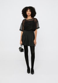 Czarna przezroczysta bluzka, plisowana czarna spódnica, czarne rajstopy i buty na wysokim obcasie. Modelka trzyma małą czarną torebkę.