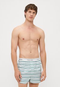 Moške bokserice v beli barvi s horizontalnimi teal črtami. Narejene so iz mehkega bombaža in imajo elastičen pas s športnim logotipom Calvin Klein.