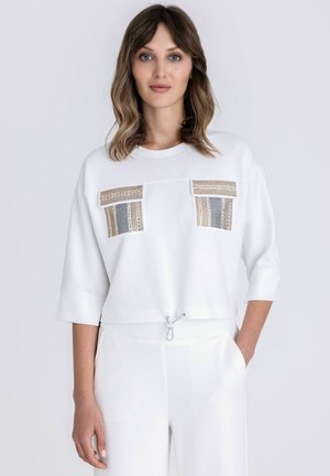 Jeune femme aux cheveux mi-longs portant un haut blanc avec des poches poitrine à motifs et un pantalon blanc assorti, debout avec une main dans la poche.