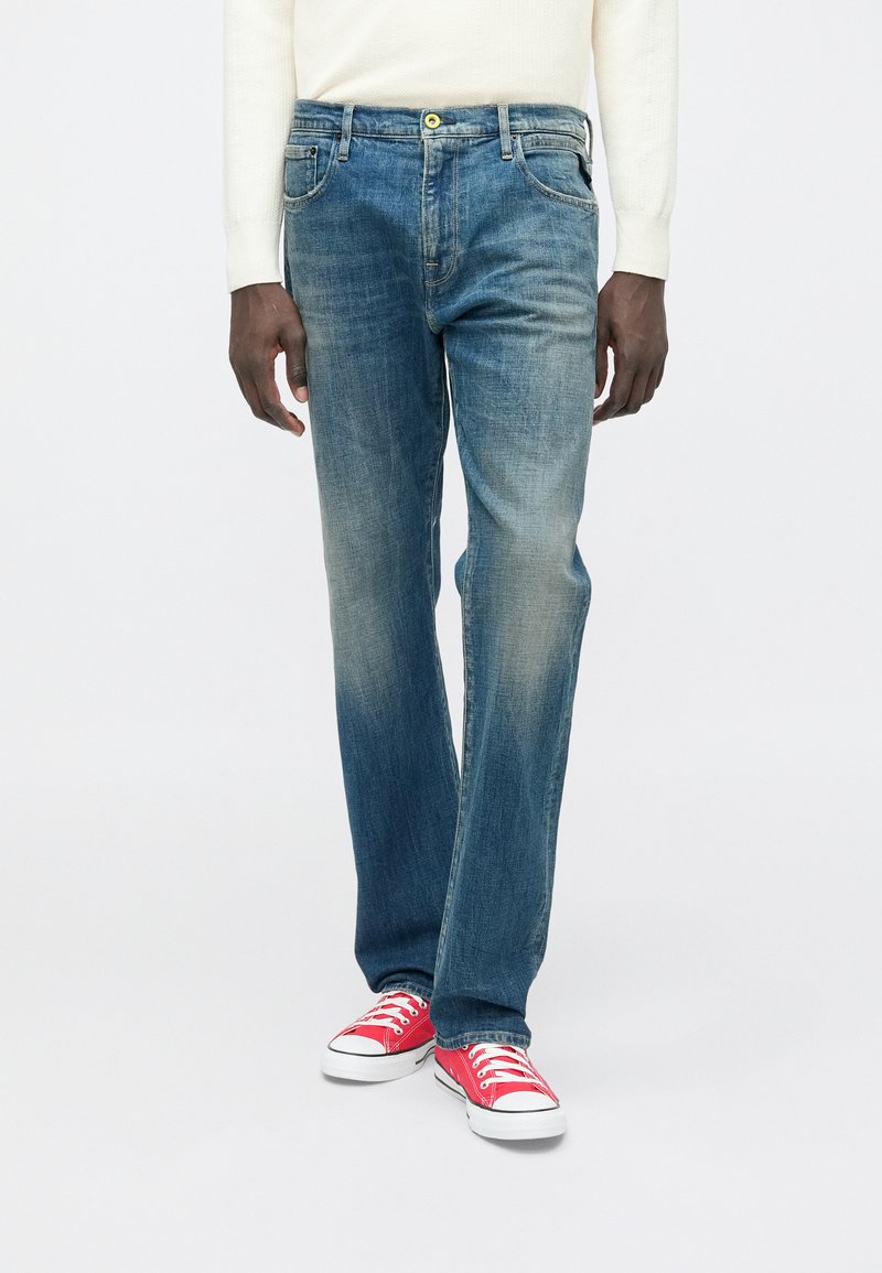 Άνδρας που φορά ξεθωριασμένο μπλε τζιν, λευκό μακρυμάνικο μπλουζάκι και κόκκινα αθλητικά παπούτσια χαμηλού ύψους, στέκεται μπροστά σε απλό φόντο.