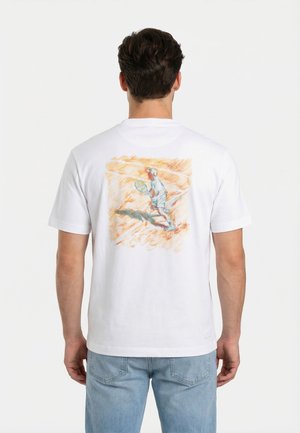 Man draagt wit T-shirt met een geschetste illustratie van een tennisser op de baan op de achterkant, gecombineerd met lichtblauwe spijkerbroek.