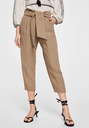 Kobieta ubrana w skrócone, brązowe spodnie o fakturze papierowej torby z szerokim paskiem związanym w kokardę, zestawione z czarnymi sandałami na obcasie z paskami.