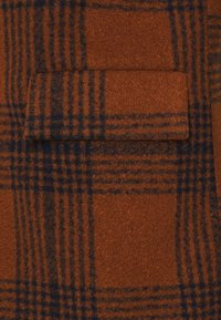 Tissu à carreaux brun avec une texture douce et des lignes bleu foncé formant un motif à carreaux. Comprend un petit détail de poche.