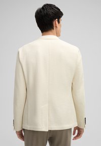 Kremowa marynarka o teksturze, z dopasowanym krojem, pojedynczym rozcięciem z tyłu i guzicznymi mankietami. Gładki materiał i minimalistyczny design, odpowiednia do eleganckiego casualu.