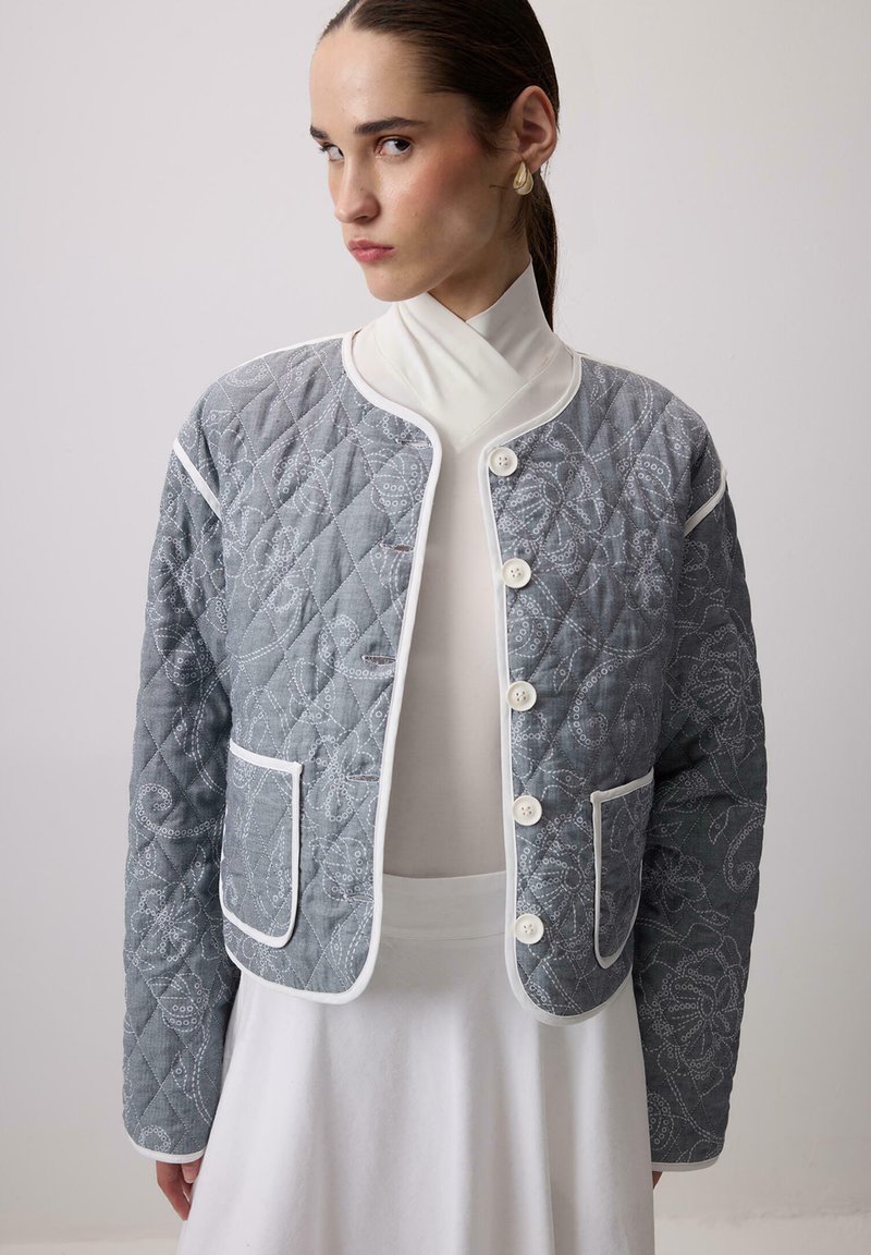 Prešívaná sivá bunda s bielymi lemami a veľkými gombíkmi, ozdobená kvetinovou výšivkou a dvoma prednými vreckami.