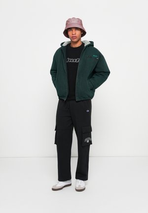 Green corduroy jacket with a fleece lining, black t-shirt with logo, black cargo pants, gray bucket hat, and white sneakers.