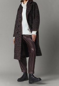 Uldblandet frakke i mørke nuancer med struktureret materiale, parret med en hvid kabelstrikket sweater og brune kunstlæderbukser, afsluttet med sorte støvler.