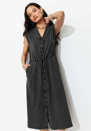 Femme aux longs cheveux foncés porte une robe midi noire sans manches boutonnée sur le devant avec des poches, accessoirisée d'un collier, d'un bracelet et de boucles d'oreilles en or.