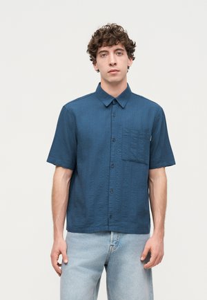 Jeune homme portant une chemise bleu marine à manches courtes avec une poche poitrine et un jean bleu clair, debout devant un fond uni.