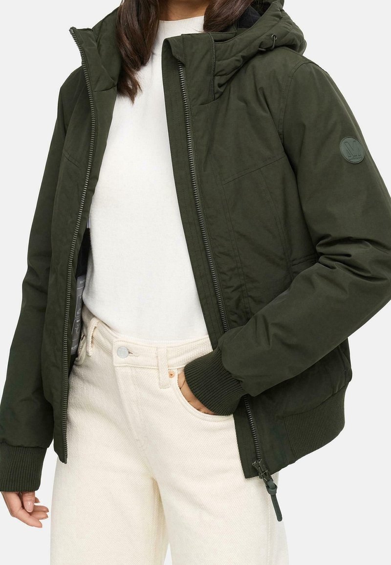 Veste zippée vert foncé avec capuche ; poignets côtelés ; écusson logo sur le bras gauche ; portée par-dessus une chemise blanche et un pantalon en velours côtelé crème.