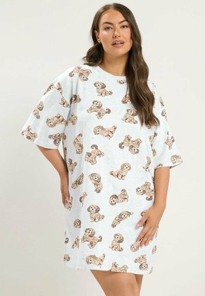 Vrouw draagt een lichtgrijze oversized jurk met bruine puppyprints, staand met één hand op haar heup, tegen een effen achtergrond.