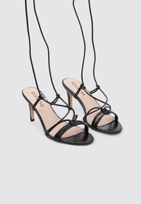 Sandalias de tacón negro con punta abierta, que cuentan con tiras delgadas, un detalle anudado en la parte frontal y una textura suave.