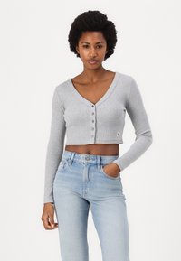 Cardigan cropped grigio a costine con maniche lunghe, scollatura a V, bottoni e piccolo logo applicato, abbinato a jeans a vita alta di colore blu chiaro.