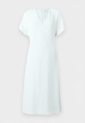 Kobieta z długimi ciemnymi włosami, ubrana w białą sukienkę midi z dekoltem w kształcie litery V i krótkimi rękawami oraz czarne wysokie sandały z piórami, stojąca.