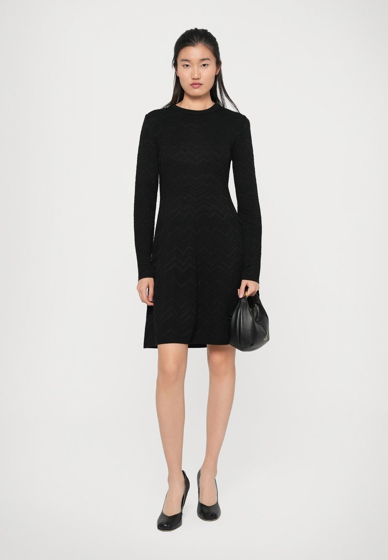 Robe noire à manches longues avec une texture en zigzag, cintrée à la taille et évasée au niveau de la jupe. Associée à des escarpins noirs et un petit sac noir.