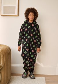 Black onesie with colorful cartoon graphics in green, red, and white. Features a zip closure and hoodie. Worn with gray clogs and patterned socks.