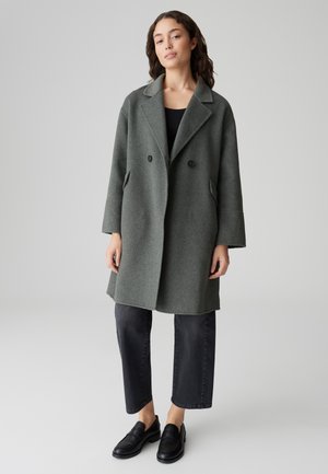 Manteau en laine gris à double boutonnage avec col cranté, poches latérales et coupe ample, associé à un pantalon large noir et des mocassins.