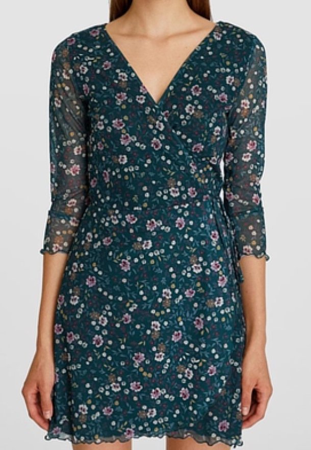 Femme portant une robe portefeuille fleurie turquoise avec des manches trois-quarts transparentes et un décolleté en V.