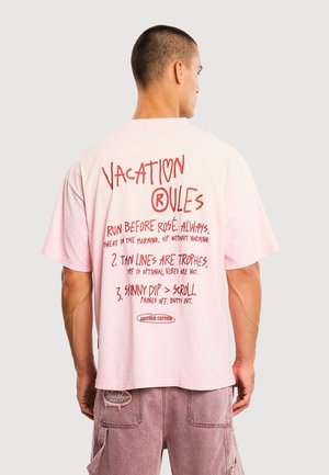 Mann trägt ein hellrosa T-Shirt mit rotem Text "Vacation Rules" und lässige mauvefarbene Shorts, von hinten gegen einen schlichten hellen Hintergrund gesehen.