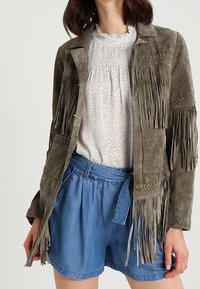 Femme portant une veste en suède marron à franges, un blouse blanche à pois, et un short bleu taille haute avec ceinture à nouer, se tenant devant un fond uni.
