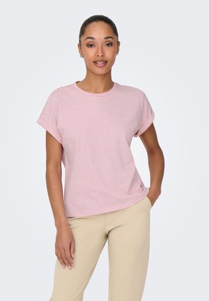 Vrouw met haar in een knot, die kleine oorringen draagt, een roze-wit gestreept T-shirt met korte mouwen en beige broek, staand met één hand in de zak.