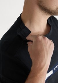 Crna majica kratkih rukava s okruglim izrezom, s detaljem gumba na ovratniku i logotipom s bijelim i plavim akcentima.