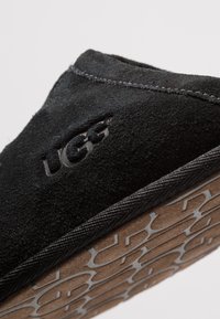 Svarte semskede tøfler med UGG-merke, beige gummisåle med teksturert mønster, og synlig søm langs kantene.