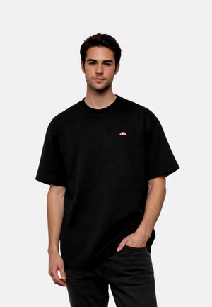 Young man with dark hair wearing a plain black oversized T-shirt and black pants, one hand in pocket, against plain light background.