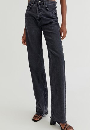 Person iført sorte jeans med høj talje, lige ben og rå kanter samt sorte hæl-sandaler med stropper mod en ensfarvet baggrund.