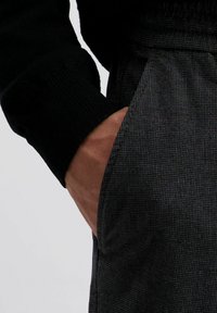 Zwarte trui met ribgebreide manchetten, gecombineerd met gestructureerde donkergrijze broek met een zak, die een subtiel geruit patroon laat zien.