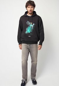 Felpa nera con stampa grafica NASA che presenta astronauti e un design cosmico color turchese. Indossata con jeans grigi chiari e sneakers nere.