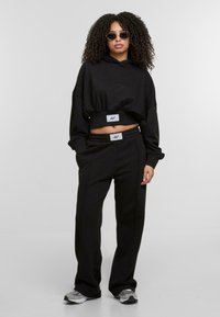 Zwarte cropped hoodie met brede mouwen, gecombineerd met zwarte high-waisted broek met een trekkoord. Beide hebben labelaccenten bij de taille.