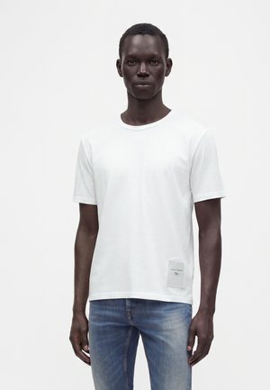 Σκούρος άντρας που φοράει απλό λευκό T-shirt με μικρό ορθογώνιο ταμπελάκι και ξεθωριασμένο τζιν μπλε, στέκεται μπροστά σε λευκό φόντο.
