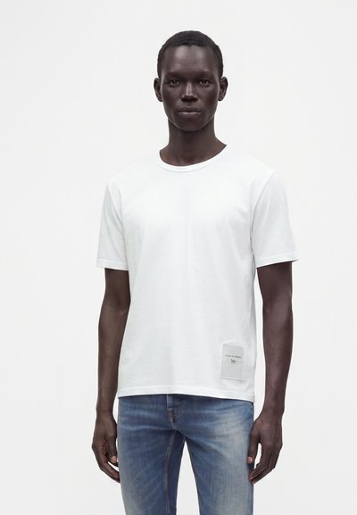 Σκούρος άντρας που φοράει απλό λευκό T-shirt με μικρό ορθογώνιο ταμπελάκι και ξεθωριασμένο τζιν μπλε, στέκεται μπροστά σε λευκό φόντο.