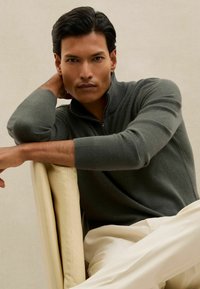 Pelēkā rāvējslēdzēja džempera ar ribotiem piedurknēm, uzlikts pār gaišiem biksēm. Gluda tekstūra, pieguļošs dizains, modelis atpūšas uz bēša krēsla.