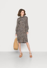 Vestido de mangas compridas, até ao joelho, com um desenho de estampado animal em bege e azul-marinho, apresentando um colarinho. Acompanhado por uma bolsa de mão em tom de areia e sandálias de salto castanhas.