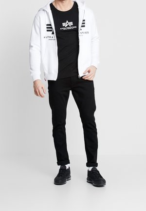 Man draagt een zwart Alpha Industries T-shirt, een witte hoodie met rits en logo, zwart opgerolde broek, witte sokken en zwarte sneakers, staand tegen een witte achtergrond.