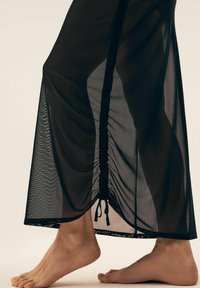 Czarna, półprzezroczysta spódnica z detalami w formie marszczenia na końcu i sznurkiem do regulacji. Posiada prosty krój oraz gładką teksturę.