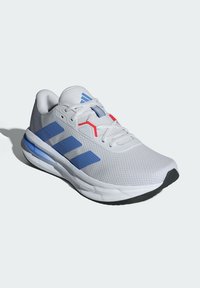 Chaussure de sport blanche avec un corps en maille texturée, accents bleus, détails rouges et semelle extérieure en caoutchouc noir. Présente trois rayures bleues sur le côté.