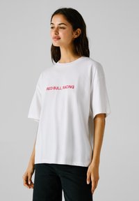 Ung kvinde iført en løs hvid t-shirt med "RED BULL RACING" i rød tekst, stående mod en ensfarvet lys baggrund.